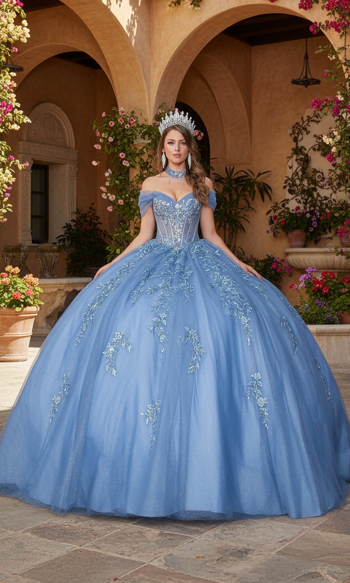 A woman in the Quinceanera Dress 5156 By Dancing Queen, featuring a sparkly blue design and sheer corset bodice, stands outdoors in a courtyard with flowers and arches, wearing a tiara.